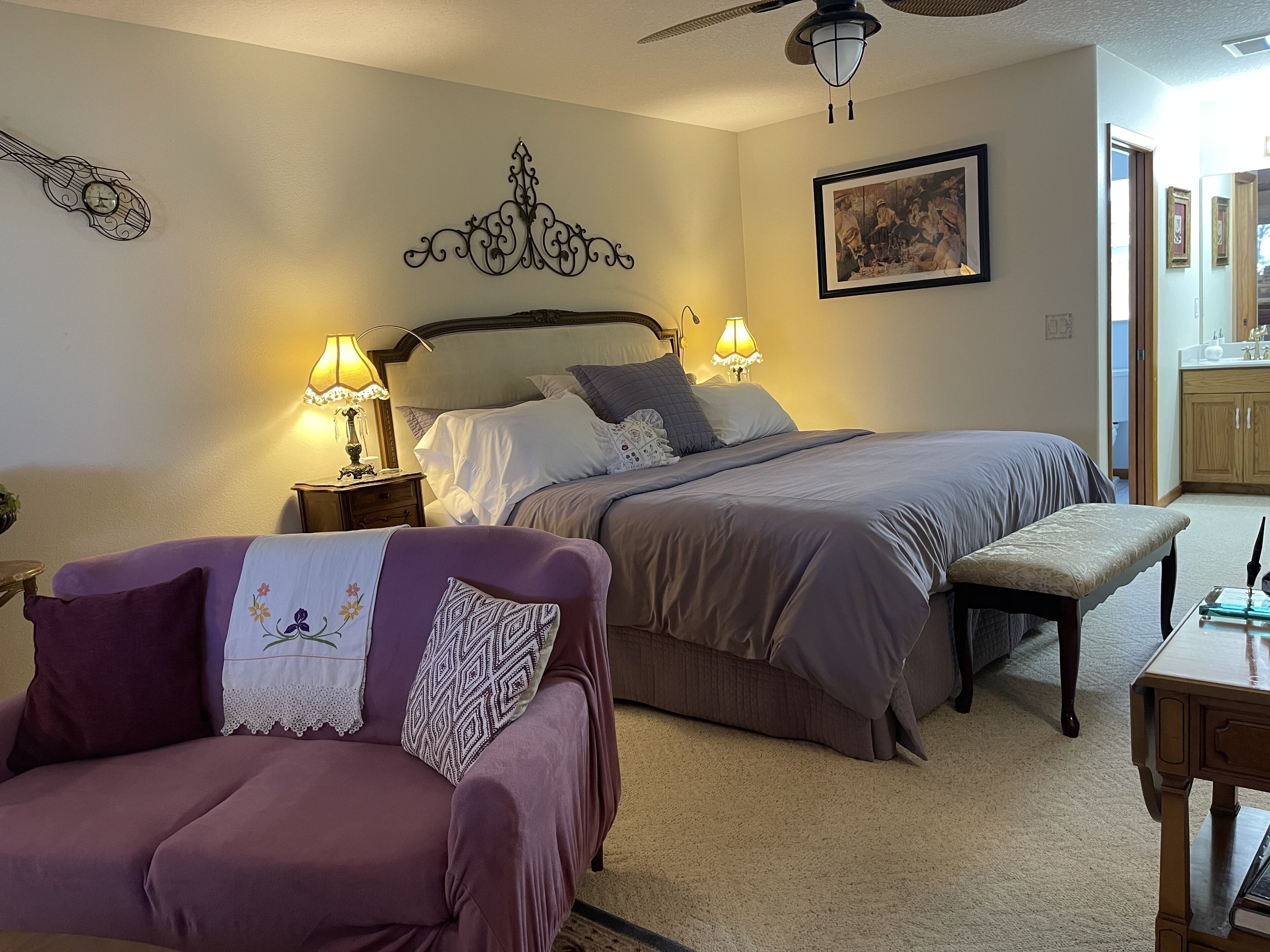 A bedroom with a bed, two bedside lamps, a ceiling fan, a chair, a bench, and a painting on the wall.