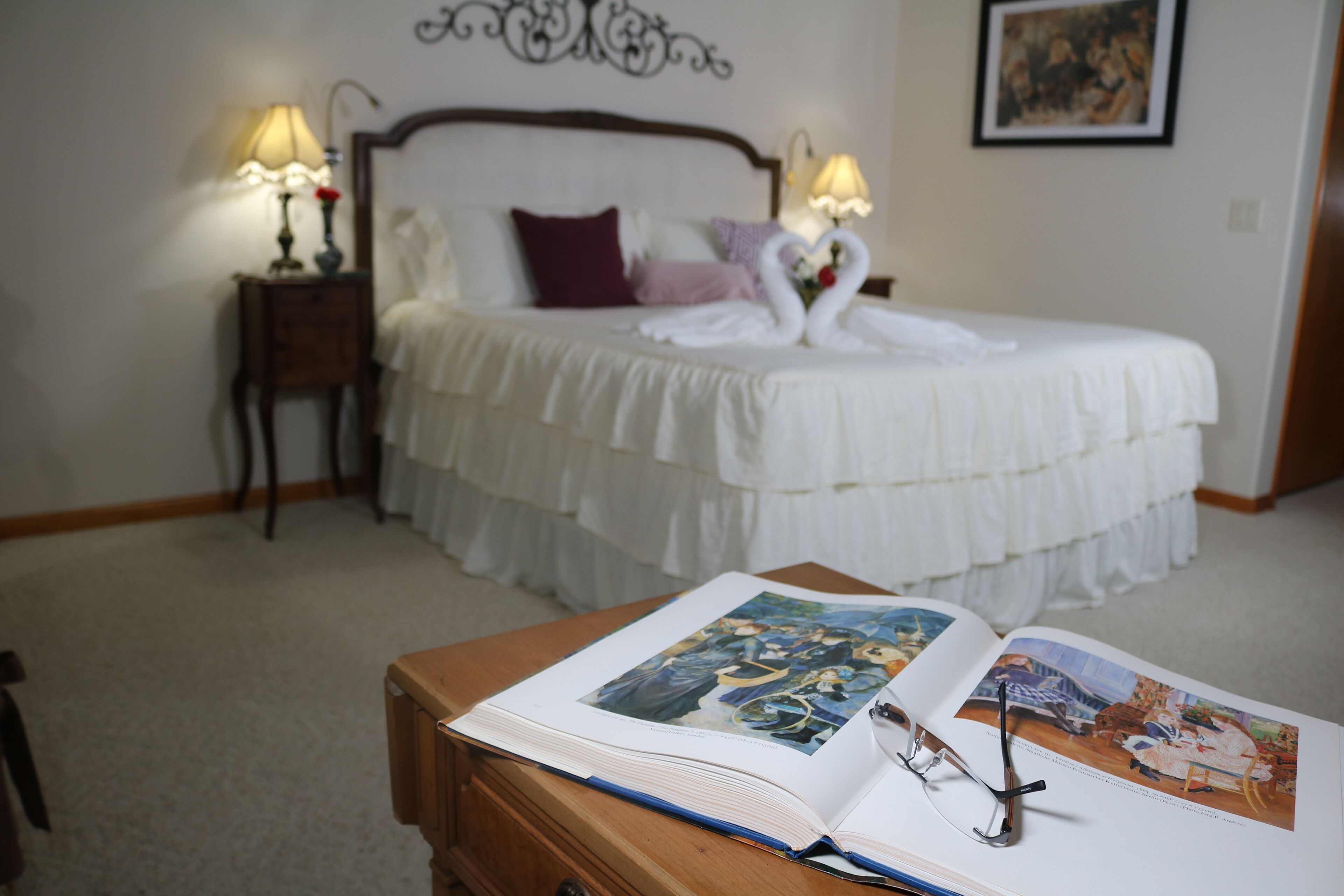 A neatly made bed with a white bedspread and pillows, a bedside table with a lamp, and a book open on a wooden dresser.