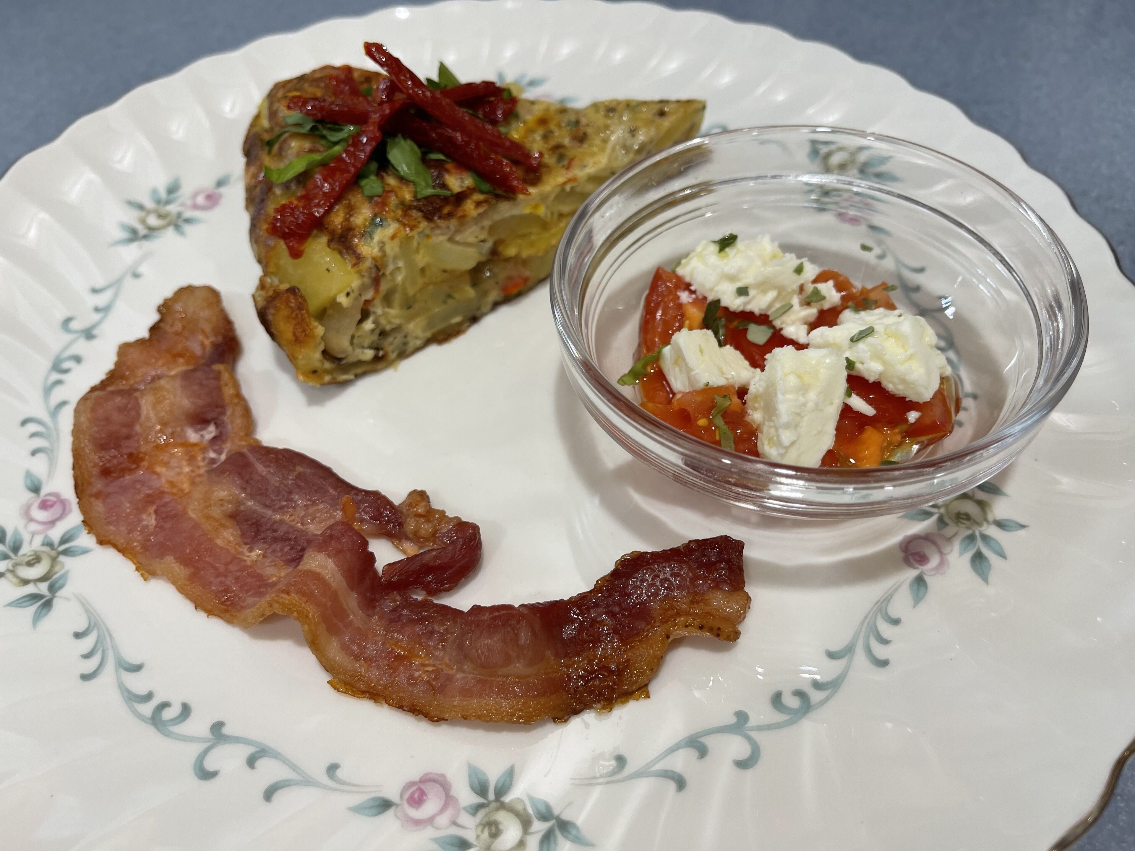 A plate with a piece of bacon, a baked potato topped with red peppers, and a bowl of tomato salad.