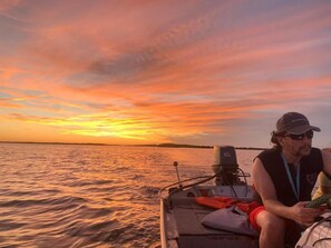 Un bateau dans lequel se trouve une personne portant une casquette et des lunettes de soleil, un gilet de sauvetage, et un objet rouge est posé sur le siège.