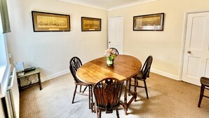 Een eetkamer met een houten tafel, vier stoelen en een bloemenvaas. Drie ingekaderde foto's hangen aan de muur en er is een deur op de achtergrond te zien.