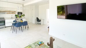 A modern kitchen with white cabinets, a dining area with a glass table and blue chairs, a flat-screen TV mounted on the wall, and a ceiling fan. - Oceanview, Minigolf, Pool, 2BR & Office. 5 min to Coco Beach (Coco)