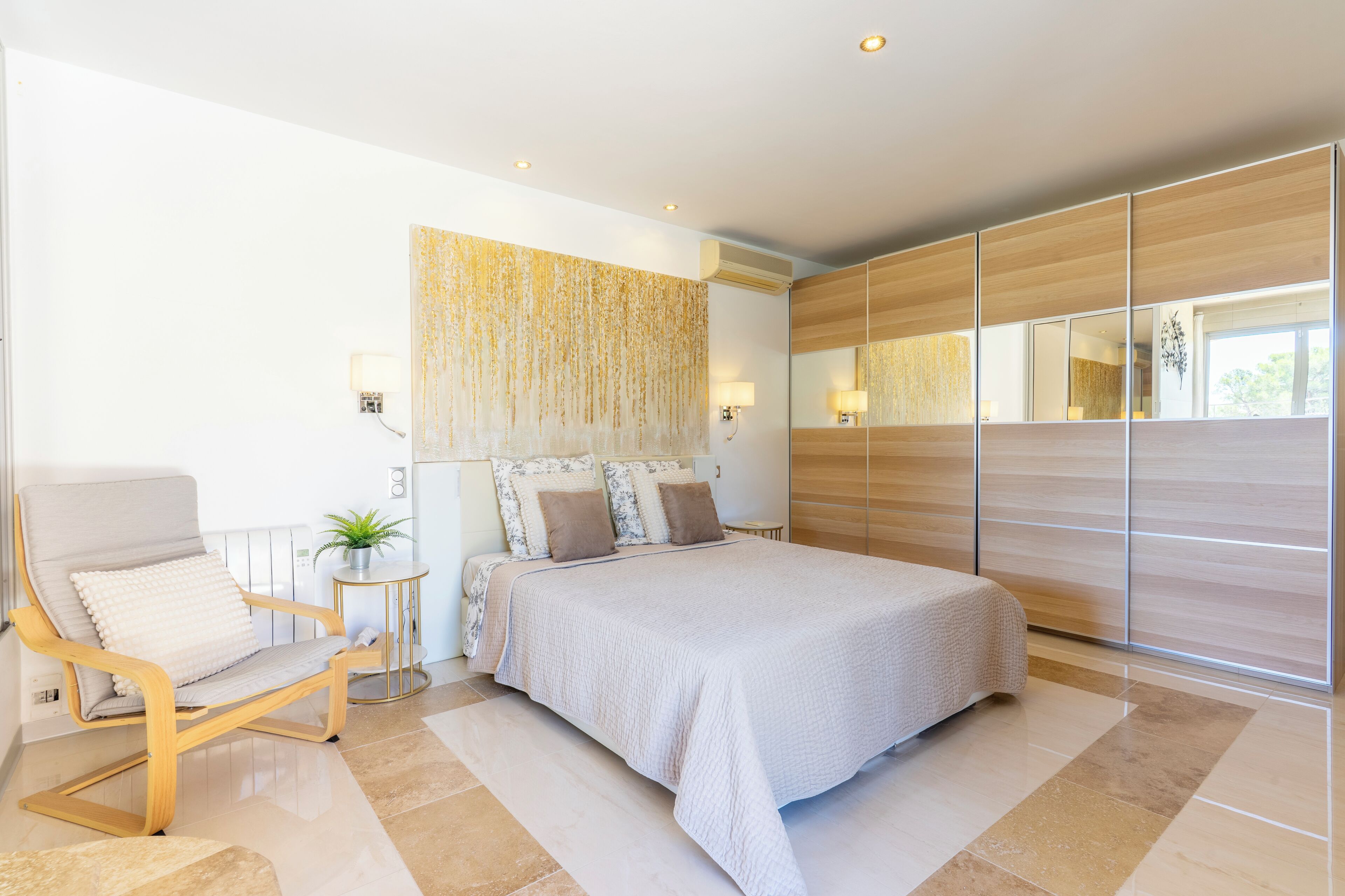 A modern bedroom with a large bed, a seating area with a chair, a small side table with a plant, and a large mirror.