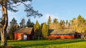 Een rustieke blokhut met een rode gevel en een metalen dak, omgeven door een weelderig groen veld en hoge bomen.