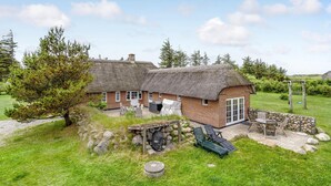 Une maisonnette rustique à toit de chaume, avec des fondations en pierre, un petit espace extérieur aménagé pour s’asseoir et un mur en pierre.
