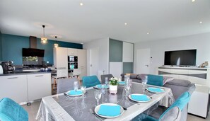 A modern dining area with a marble table set for six, surrounded by teal chairs. A kitchen with white cabinets and a black stove is visible in the background. - La Villa de L'océan - Erdeven (ERDEVEN)