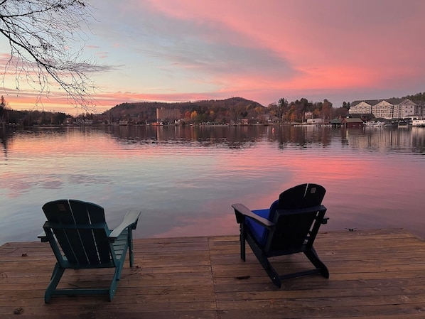 Two Adirondack chairs on a wooden dock overlooking a calm lake with a sunset in the background.