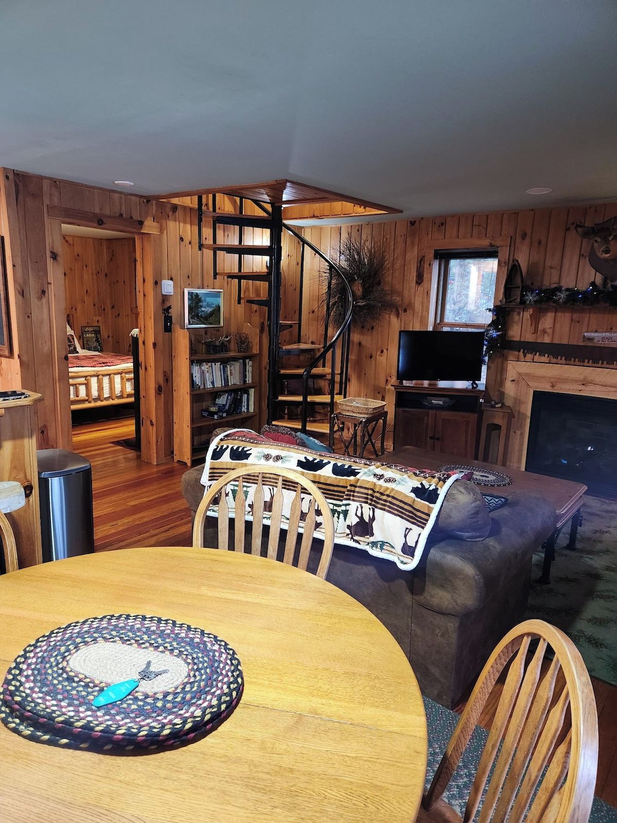 A cozy cabin interior with a dining table, chairs, a fireplace, a television, and a spiral staircase.