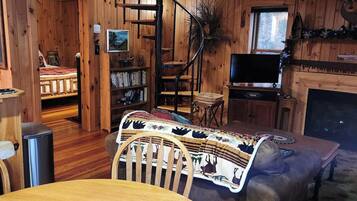 A cozy cabin interior with a dining table, chairs, a fireplace, a television, and a spiral staircase.