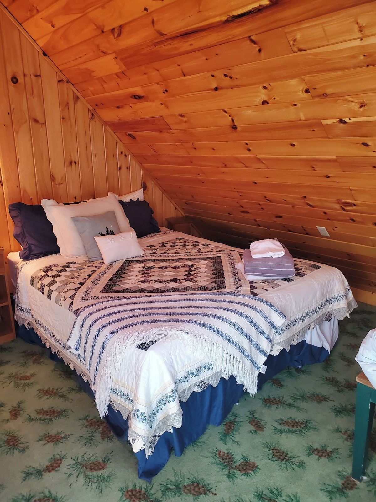 Ein Schlafzimmer mit schrägverlegtem Holzboden, einem Bett mit einem gemusterten Bettdeckbett und einem grünen floralen Teppich.