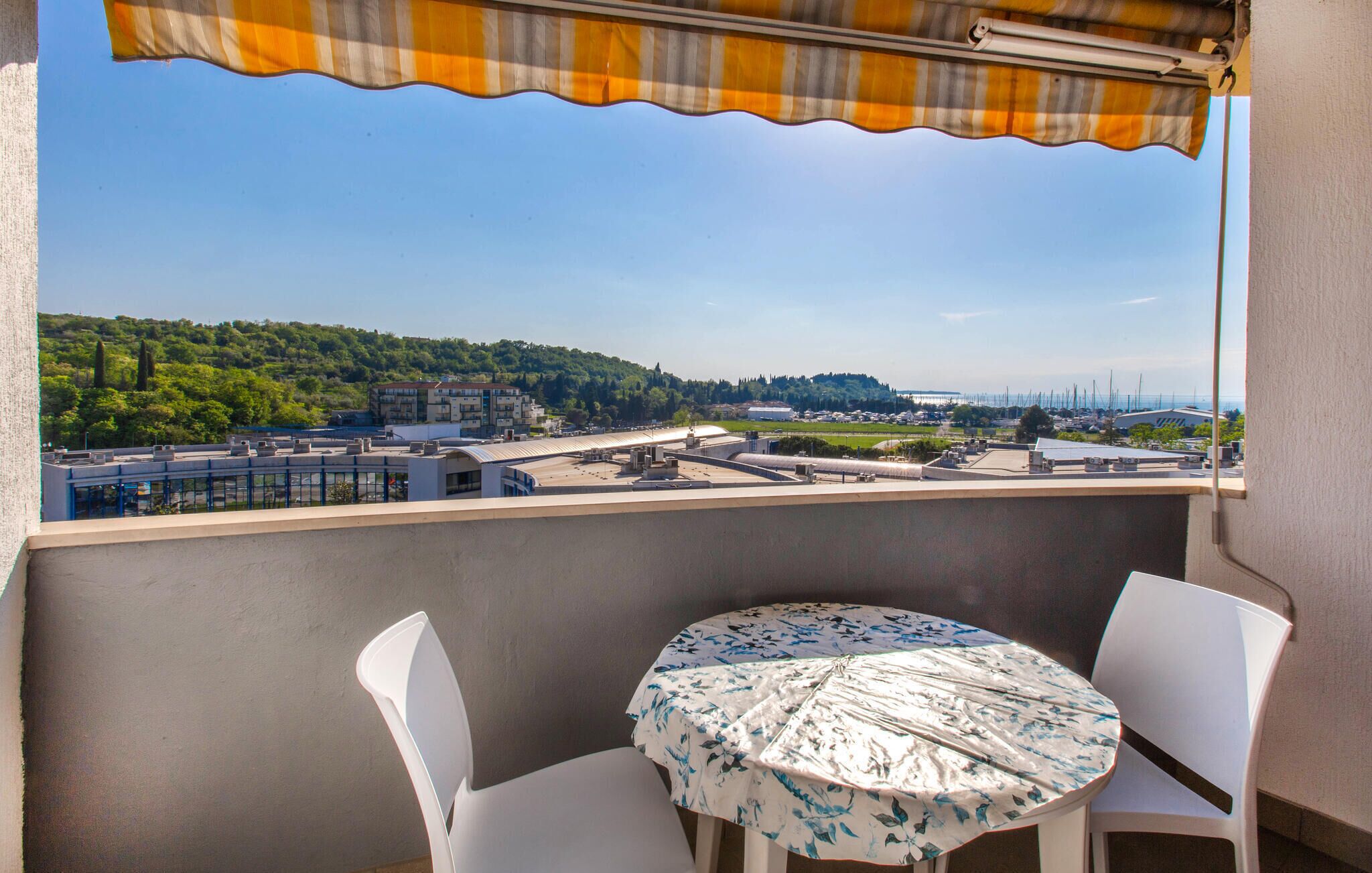 En balkon med et bord og stole, udsigt over bybilleder med bygninger og et fjernt hav.