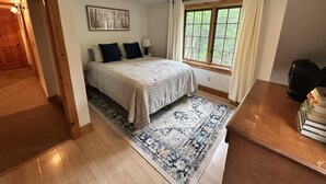 Un dormitorio con cama, ventana con cortinas, alfombra y mesita de noche.