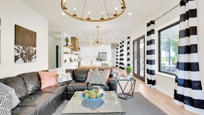 Un salon moderne avec un canapé en cuir, une table basse en verre sur laquelle sont disposés des fruits, et une vue sur la cuisine et la salle à manger.