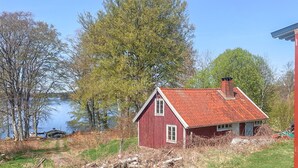 Ett rött trähus med ett rött tak vid en sjö, omgivet av träd och en klar himmel.