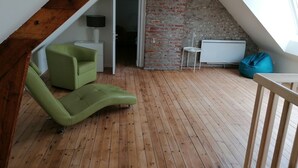 Un dormitorio tipo loft con techo inclinado, piso de madera, una silla verde, una mesita, un sillón de peluche azul y una pared de ladrillo.