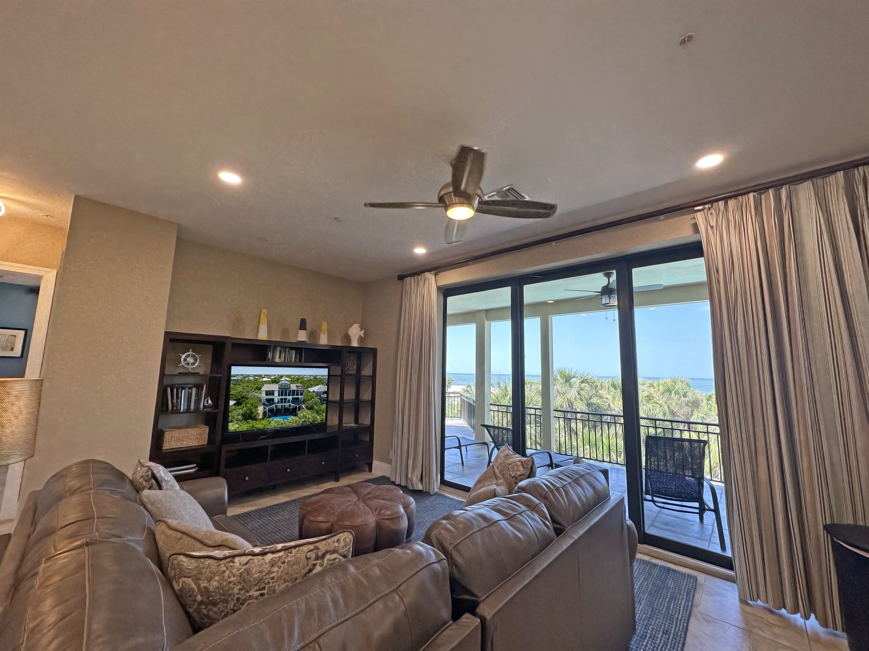 A living room with a leather sofa, a TV on a wooden stand, and a balcony with a view of palm trees and the ocean.