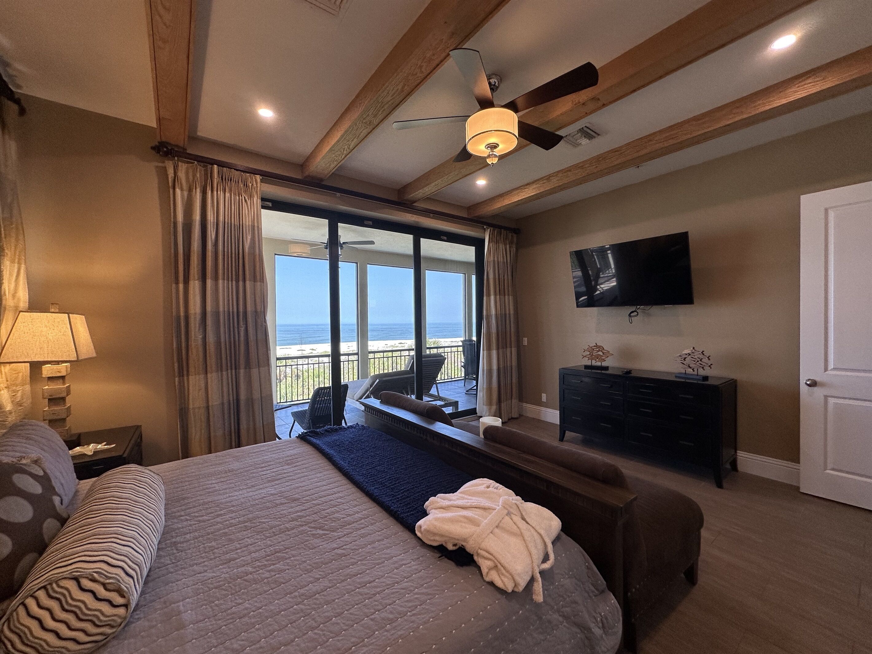 Un dormitorio con cama, mesita de noche, lámpara, cómoda, televisor y balcón con vista al océano.