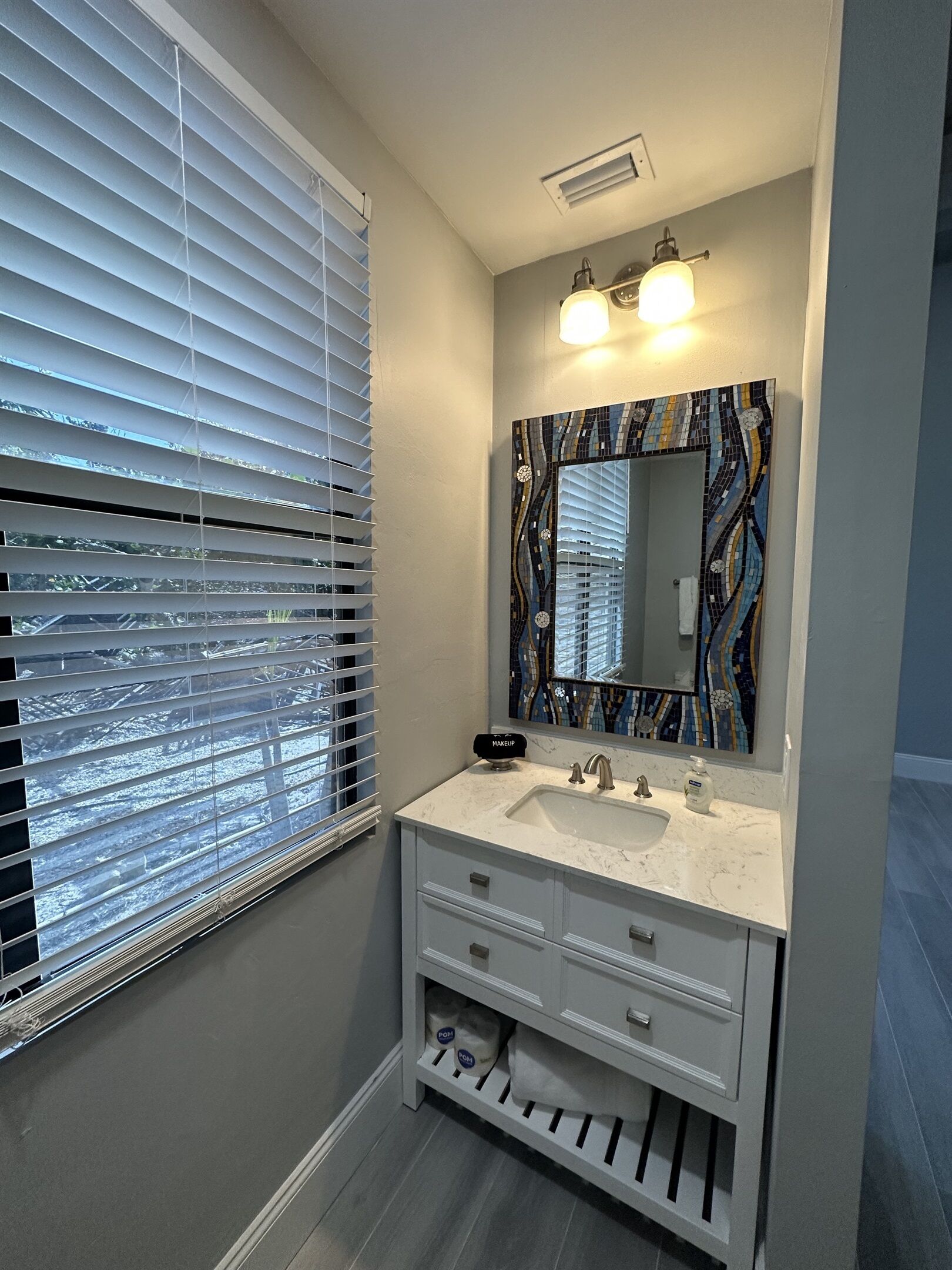 A bathroom with a white vanity, a mirror with a mosaic backsplash, and a window with blinds.