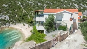 A two-story house with a red-tiled roof, located near a beach with clear turquoise water.