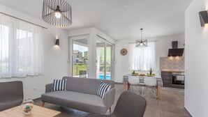A modern living room with a sofa, a dining table with chairs, a kitchen area with a stove, and a view of the outside through a glass door. - Awesome apartment in Vrbnik with WiFi (Vrbnik)