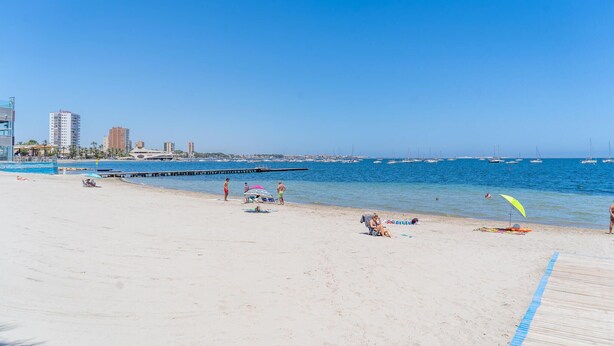 Una playa con cielos despejados, mar tranquilo y edificios al fondo.