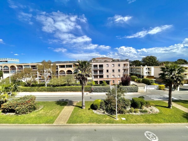 A multi-story building with balconies, surrounded by well-maintained greenery and a clear blue sky.