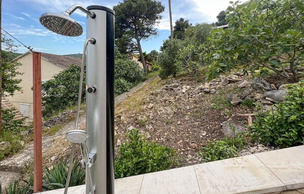 A metallic outdoor shower with a hose attachment, set against a backdrop of a rocky garden and a residential building. - Beach front home in Splitska with house sea view (Splitska)