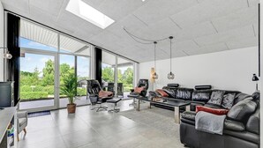 A spacious living room with a large black leather sofa, a coffee table, and a dining table. There are two chairs and a potted plant. The room has large windows and a view of greenery outside.