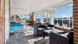 A covered outdoor area with a swimming pool, wicker furniture, and large windows offering views of the landscape. - Beautiful home in Ringkøbing with sauna (Ringkøbing)