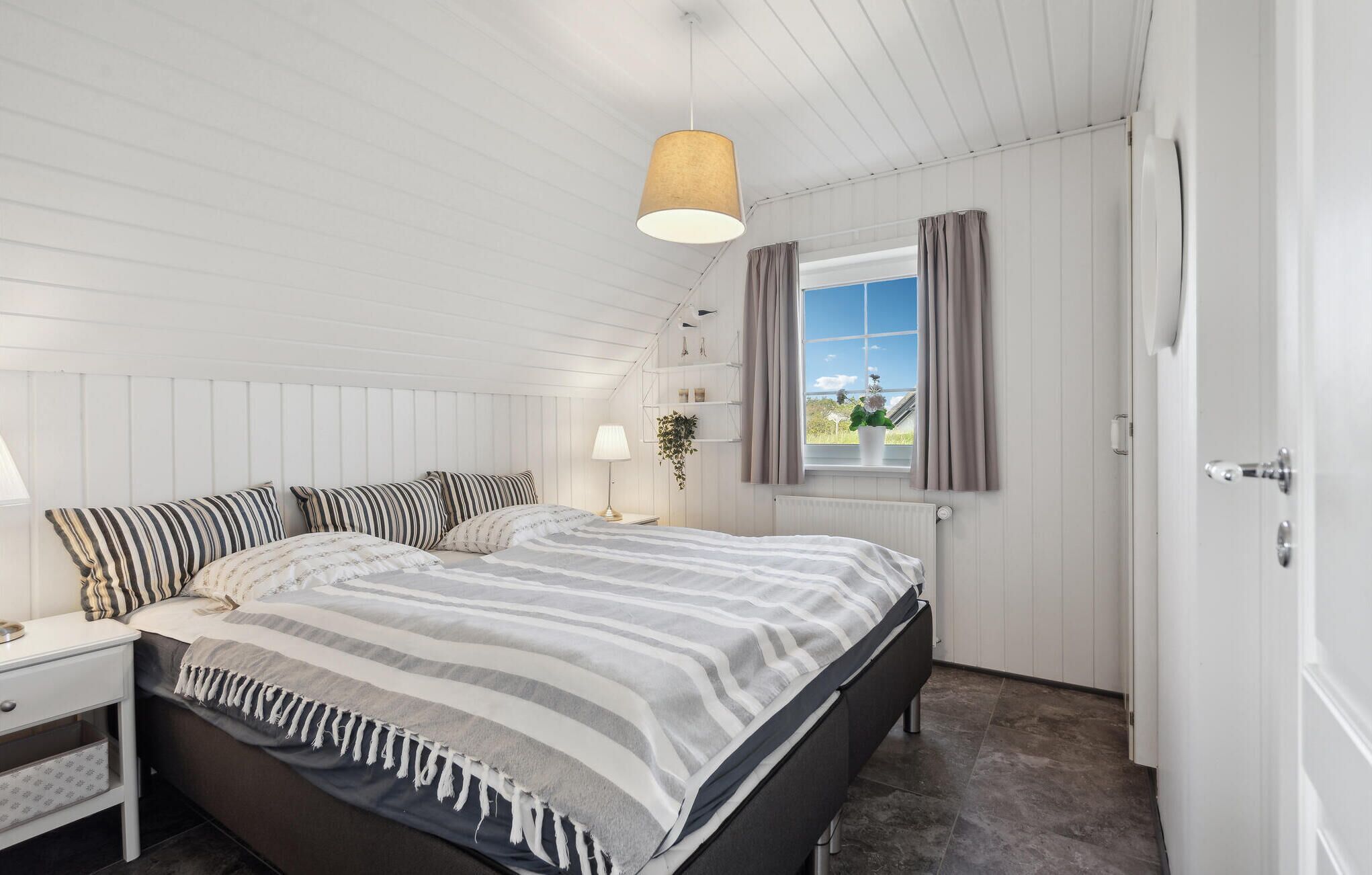 Un dormitorio con cama, mesitas de noche, lámpara colgante, ventana con cortinas y vista al exterior a zonas verdes.