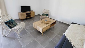Una habitación con una mesa de centro de madera, dos sillas de mimbre, un televisor sobre un mueble de madera y una pared blanca con una lámpara colgante.