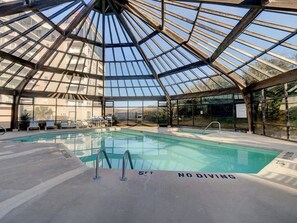 Une piscine intérieure, protégée par des parois en verre, avec un panneau indiquant qu’il est interdit de plonger, des chaises longues et une vue sur les bâtiments environnants.