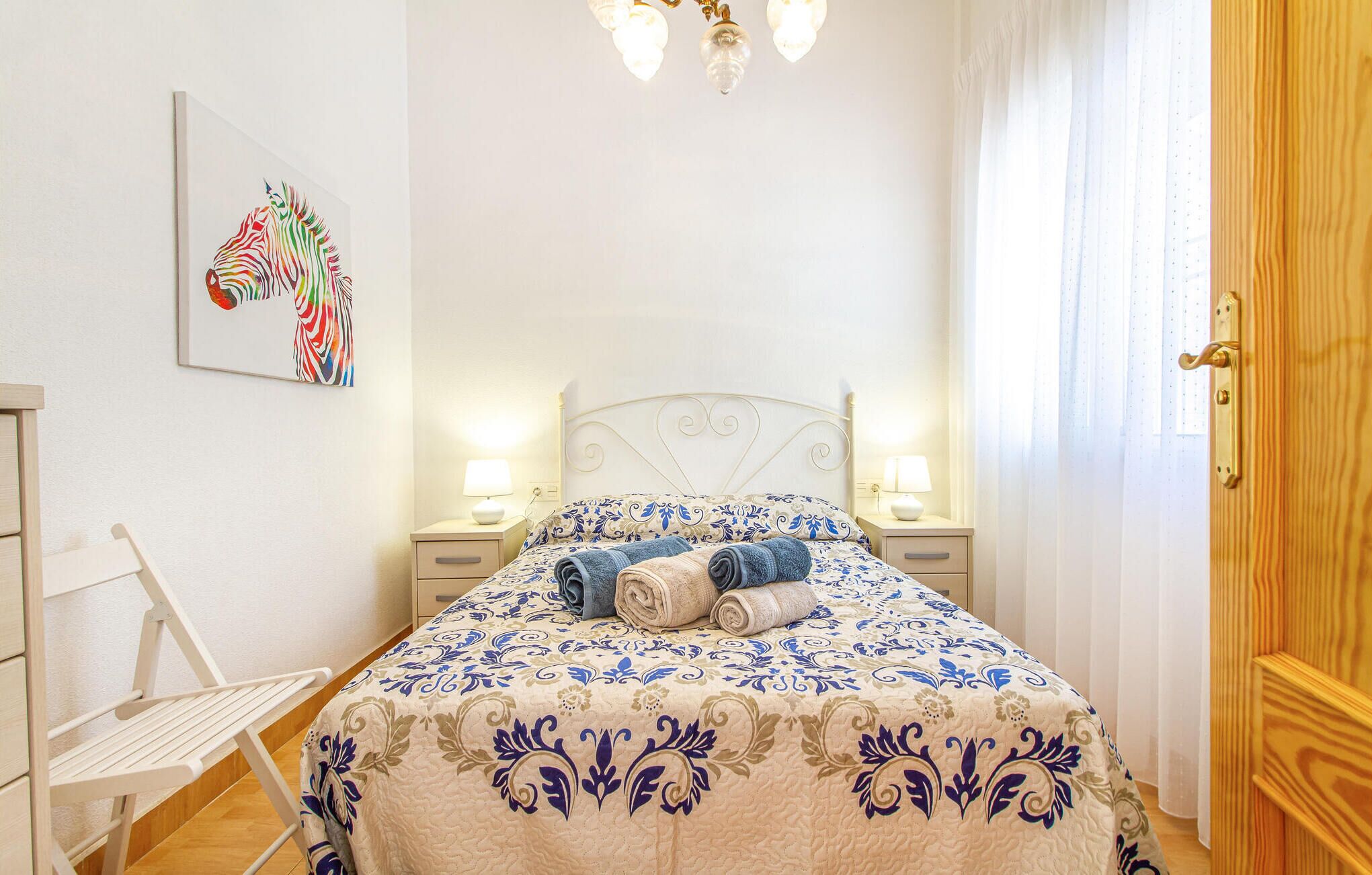 A neatly made bed with a patterned bedspread, two bedside lamps, a wall-mounted artwork, and a wooden door.
