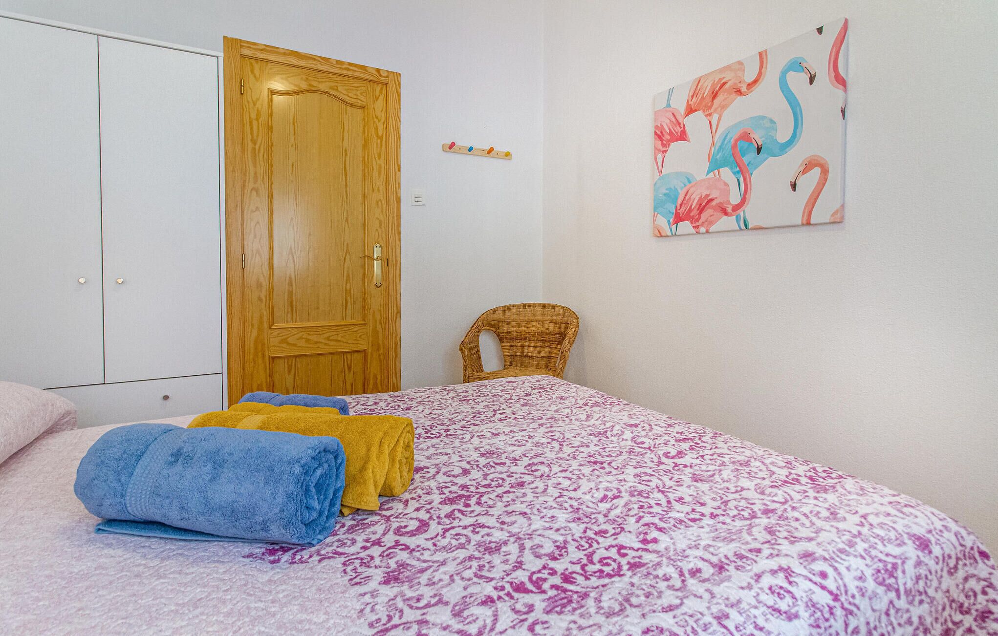 Ein Schlafzimmer mit einem Bett, einer Holztür, einem Korbstuhl und einer Wand mit einem Flamingo-Gemälde.