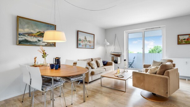 A modern living room with a wooden dining table, a sofa, a coffee table, and a view of the outdoors through a sliding glass door. - Cozy apartment in Gudhjem (Gudhjem)