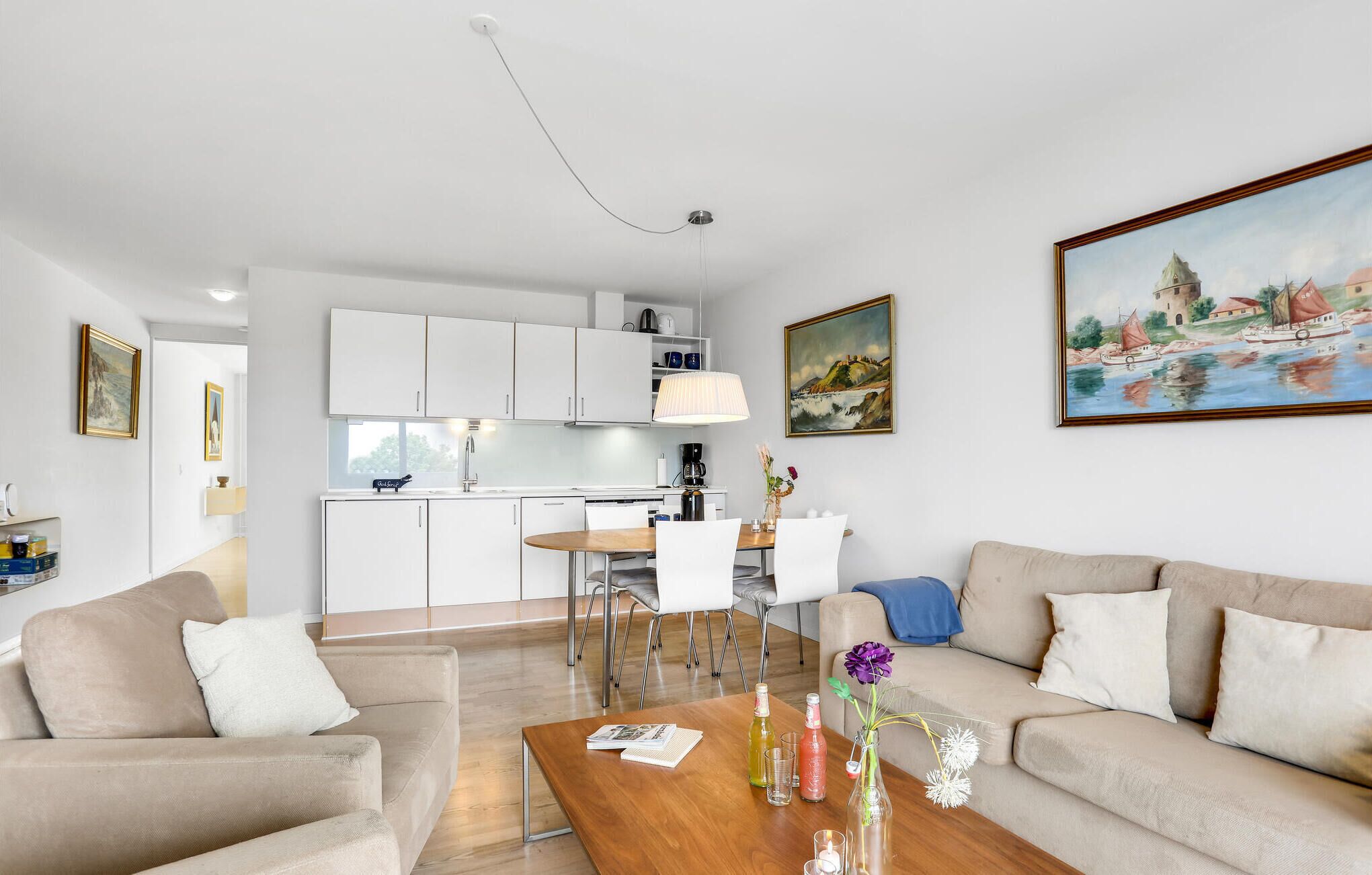 A modern living room with a sofa, coffee table, and a painting. There is a kitchen area with cabinets and a dining table.