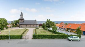 A church with a tall steeple, a paved driveway, and a parking area with cars. - Cozy apartment in Gudhjem (Gudhjem)