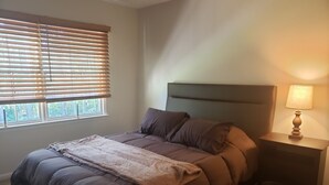A bedroom with a bed, a nightstand with a lamp, a window with blinds, and a view of trees outside. - Comfortable 3 Bedroom, 3 Bathroom With Amenities! (Savannah)