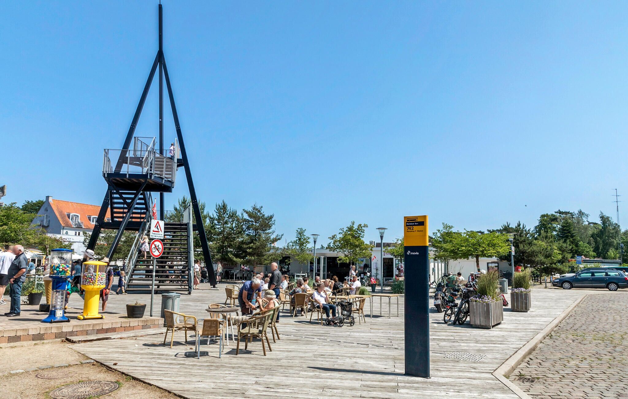 Une place publique avec des sièges en plein air, un pont piétonnier et un panneau indiquant un arrêt de tramway.