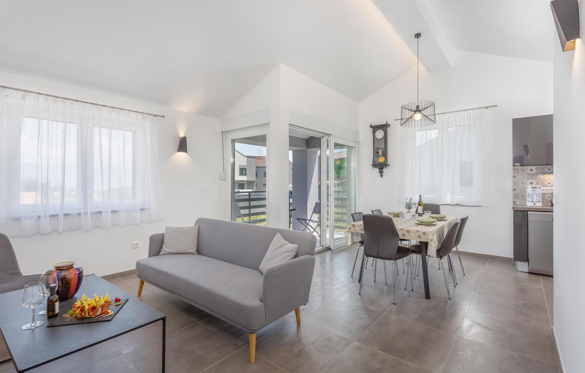 A modern living room with a grey sofa, dining table with chairs, and a kitchen area with a stove and cabinets.
