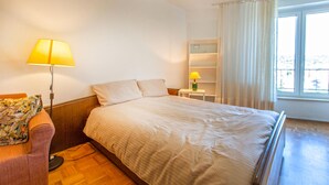 Une chambre d’hôtel avec un lit, une table de chevet avec une lampe, une chaise, une petite étagère et une fenêtre avec des rideaux.