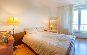 A hotel room with a bed, a bedside table with a lamp, a chair, a small shelf, and a window with curtains. - Beautiful apartment in Portoroz (Portoroz)