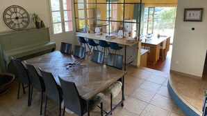 Ein Essbereich mit großem Holztisch und Stühlen, ein Küchenbereich mit Theke und Sitzgelegenheiten sowie ein Blick ins Freie durch große Fenster.