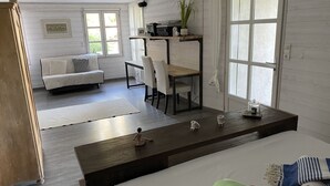 Ein modernes Zimmer mit einem Bett, einem Schreibtisch, einem Sessel und einem Sofa. Es gibt Fenster, eine Pflanze und einen kleinen Tisch mit einer Lampe.