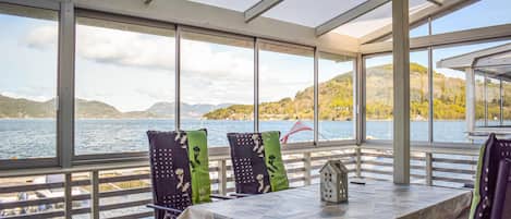A covered outdoor seating area with a table and chairs, overlooking a lake with hills in the background.