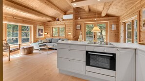 A cozy cabin interior with a wooden ceiling, large windows, a sofa, a dining area, and a kitchen with a Bosch oven. - Cozy home in Ulfborg with kitchen (Ulfborg)