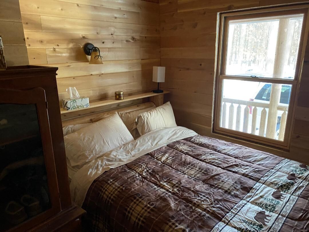 A wooden cabin room with a bed, a nightstand with a lamp, and a window with a view of a wooden deck.