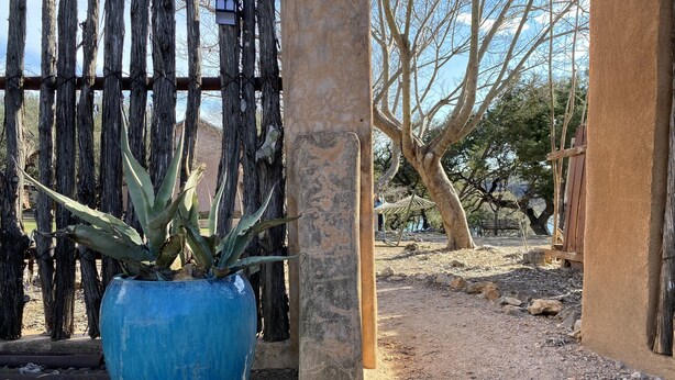 Un pot bleu contenant une plante, placé dans un décor extérieur rustique agrémenté d’une clôture en bois, d’un pilier en béton et d’un chemin de terre.