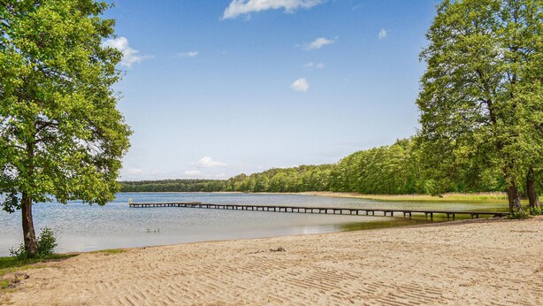 A sandy beach with a wooden pier extending into a calm lake, surrounded by dense green trees.
