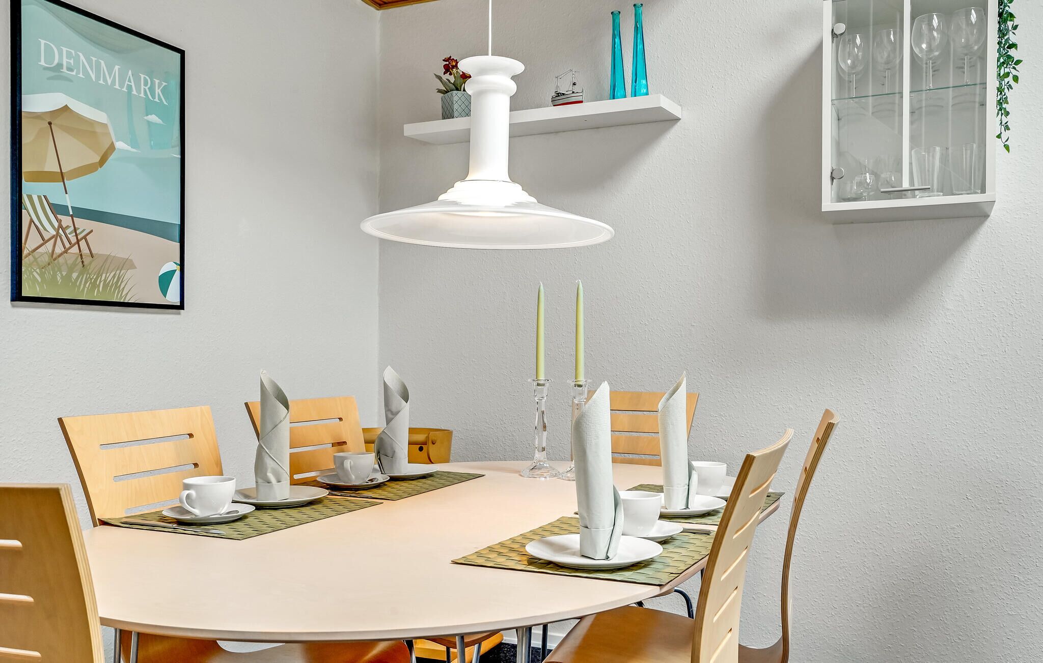 A dining area with a round table, chairs, a white pendant light, and a framed picture of a beach scene.